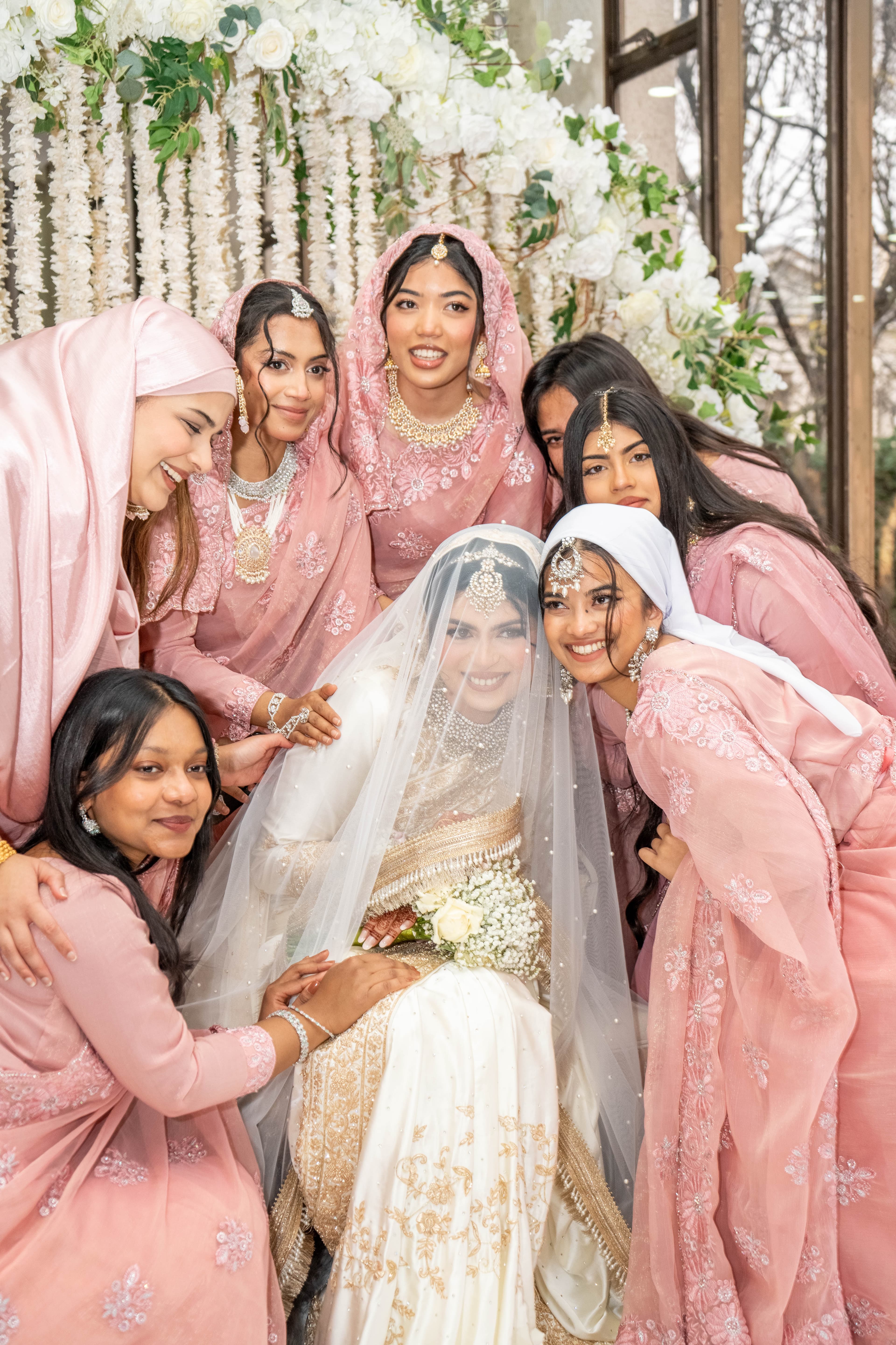 Group gathered around a seated bride beneath white floral decor.